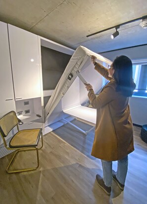 Una persona instalando un catre blanco, plegable y fijado a la pared, en una habitación con suelo de madera, una silla y un ventanal.