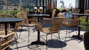 Un área de estar al aire libre con sillas y mesas de mimbre, dos vasos de cerveza y una vista de la ciudad al fondo.