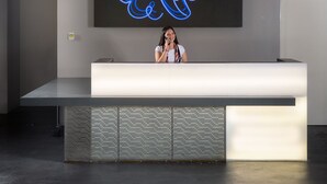 Un mostrador de recepción con una amplia pantalla digital que muestra gráficos azules abstractos, un diseño moderno con techo de metal y hormigón, y un área de recepción minimalista.