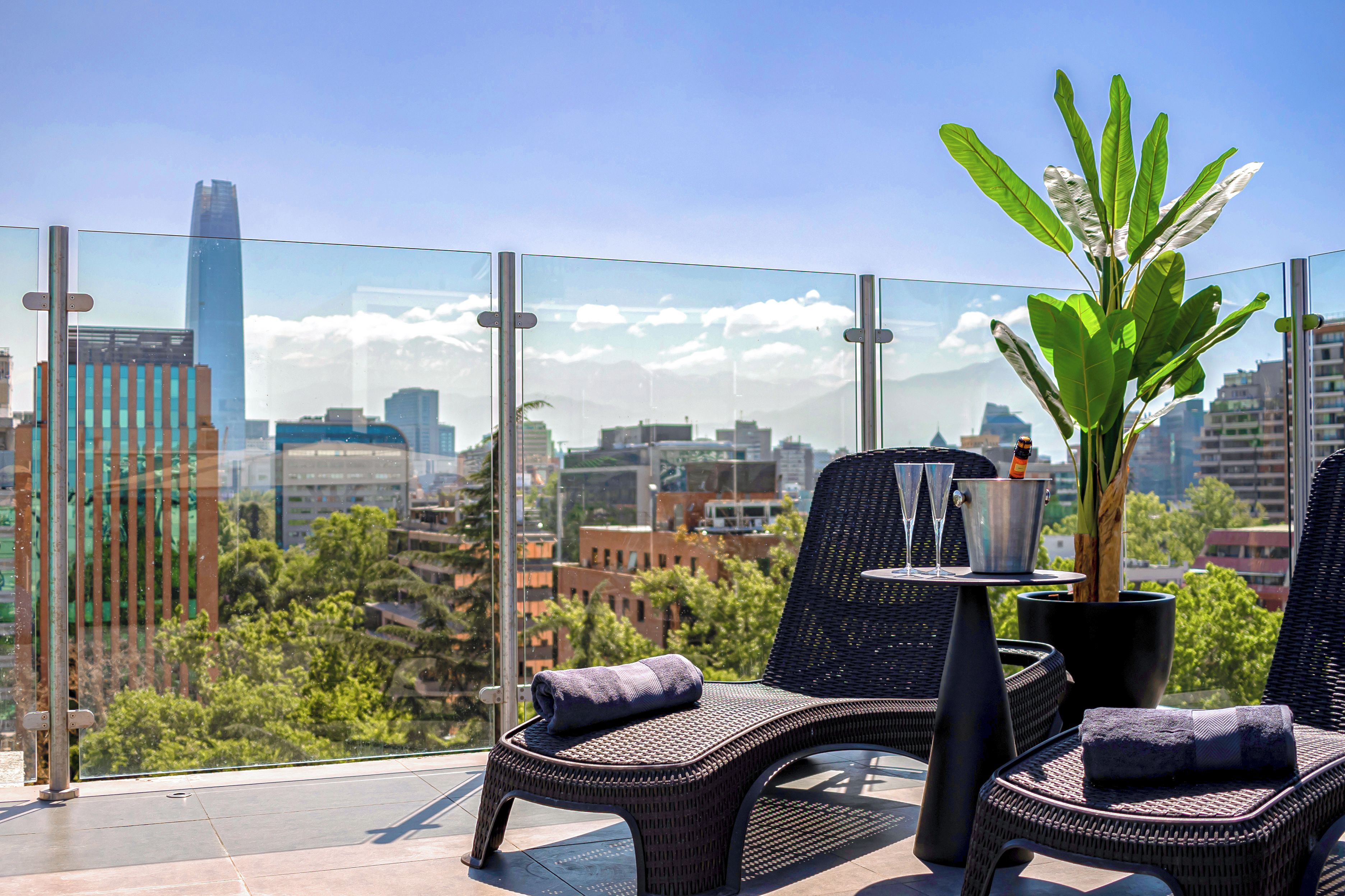 Un balcón con sillas de mimbre, una mesa, una planta en maceta y vistas a un paisaje urbano.