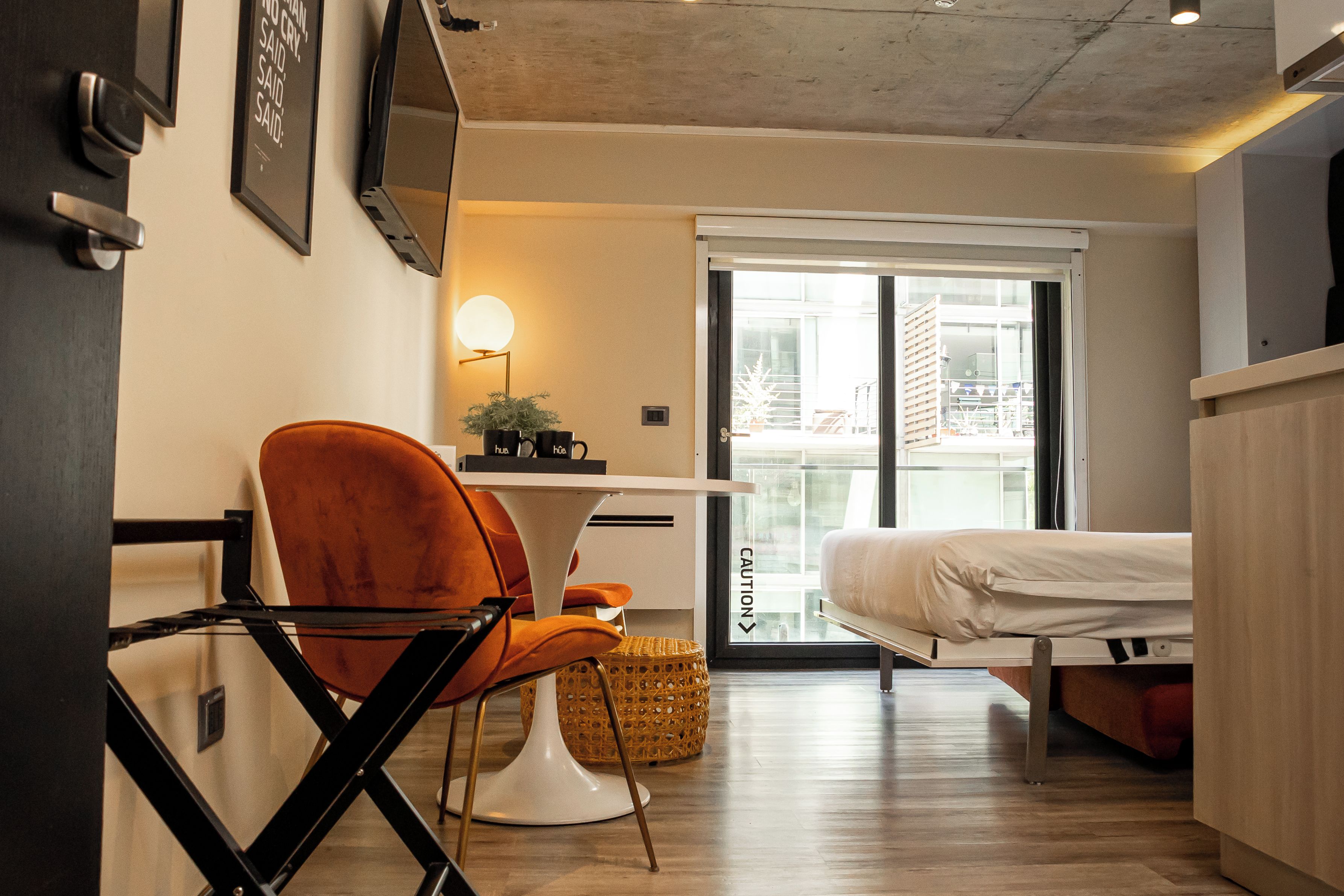 Una habitación de hotel moderna con cama, escritorio, silla, planta en maceta y puerta corrediza de vidrio que da a un balcón.
