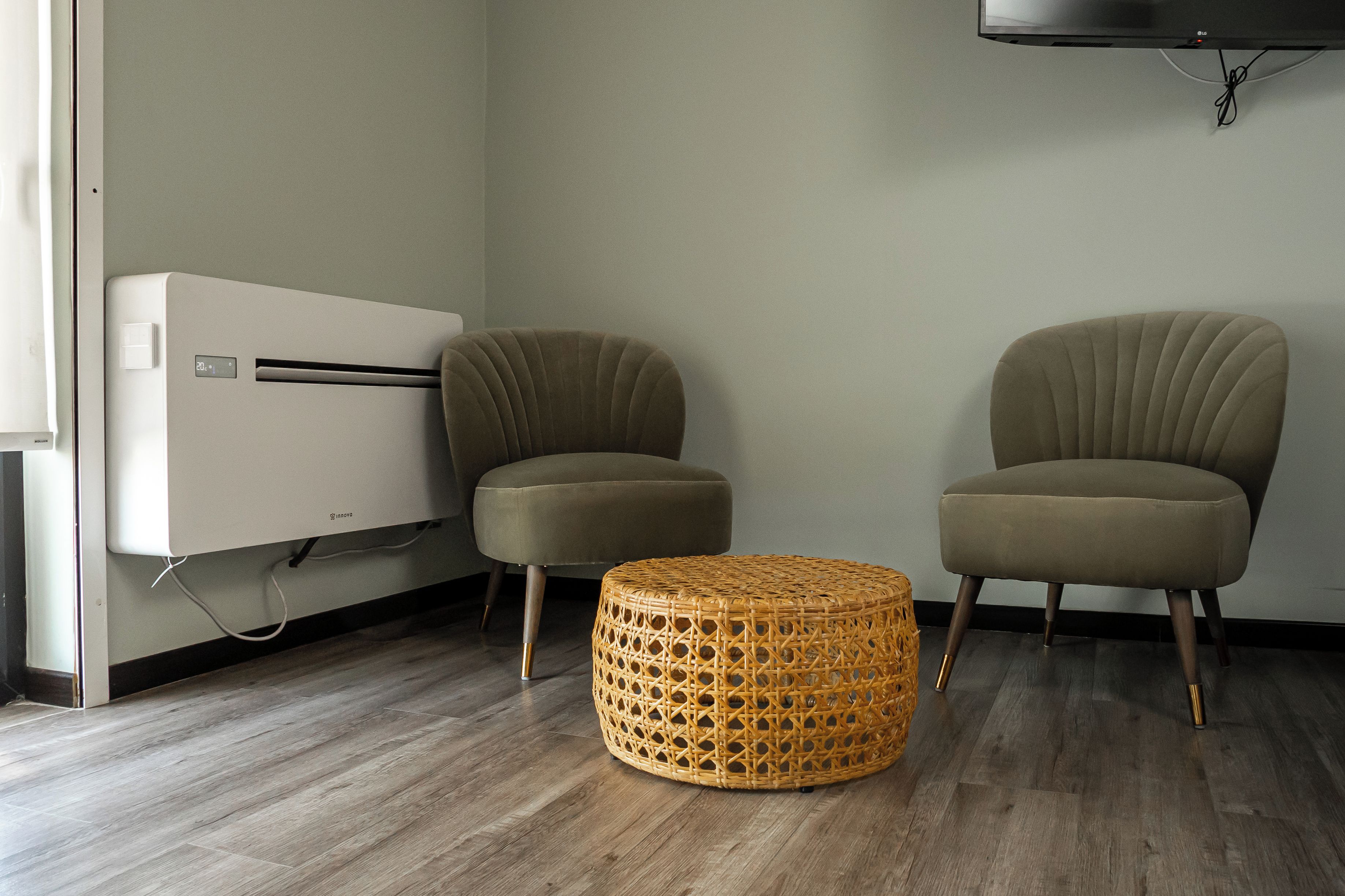 Une pièce comprenant deux fauteuils rembourrés, un pouf en osier, un climatiseur blanc et un téléviseur à écran plat fixé au mur.