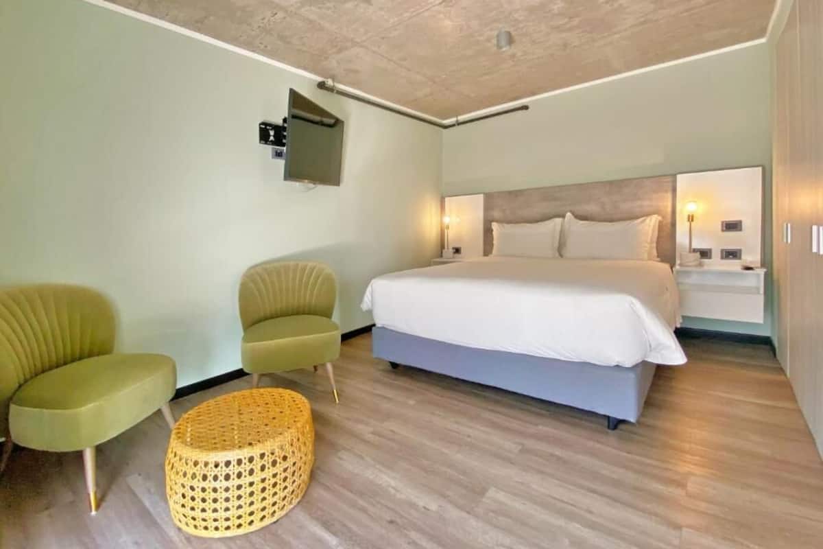 Un dormitorio moderno con una cama, dos sillas verdes, un reposapiés de mimbre y techo de madera.