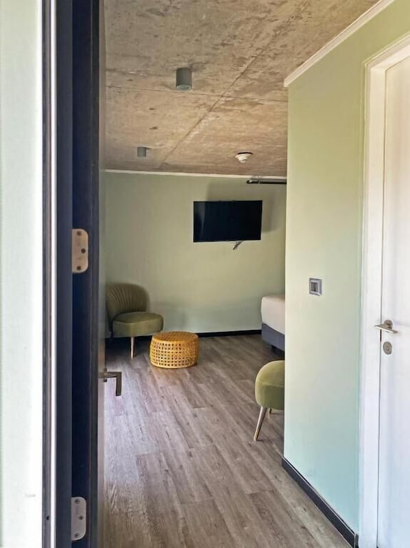 Una habitación con piso de madera, un sofá verde, un reposapiés pequeño y redondo, un televisor montado en la pared y una puerta con manija.