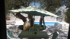 Área externa com mesas e cadeiras sob um grande guarda-sol, cercada por árvores e vista para uma piscina.