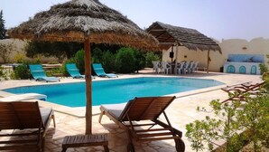 Uma área de piscina com espreguiçadeiras, um guarda-sol de palha e uma mesa de jantar com cadeiras.