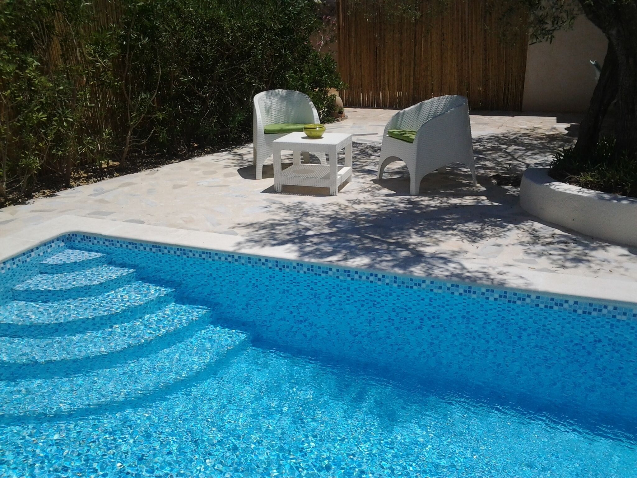 Zwembad met twee witte stoelen en een kleine tafel, omgeven door een betonnen terras en weelderige begroeiing.