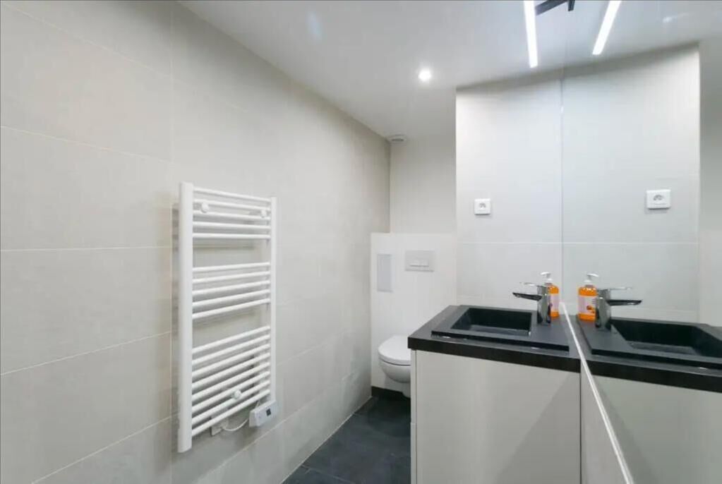 A modern bathroom with a white tiled wall, a white radiator, a toilet, and a black sink with two soap dispensers.
