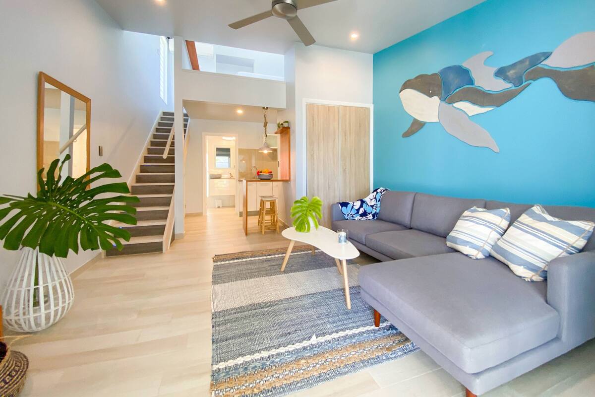 A modern living room with a grey sofa, a blue mural, a staircase leading to an upper level, and a kitchen in the background.