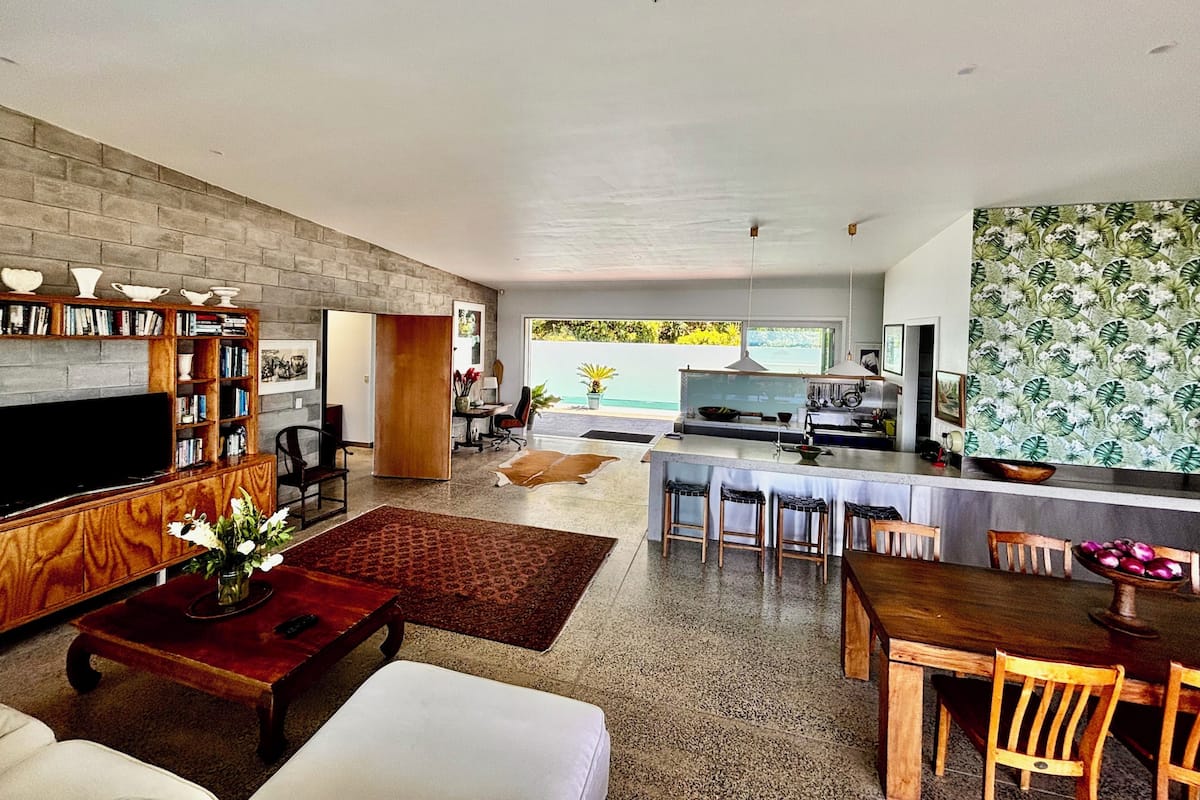 A modern living room with a flat-screen TV, a wooden bookshelf, and a dining area with a kitchen in the background.
