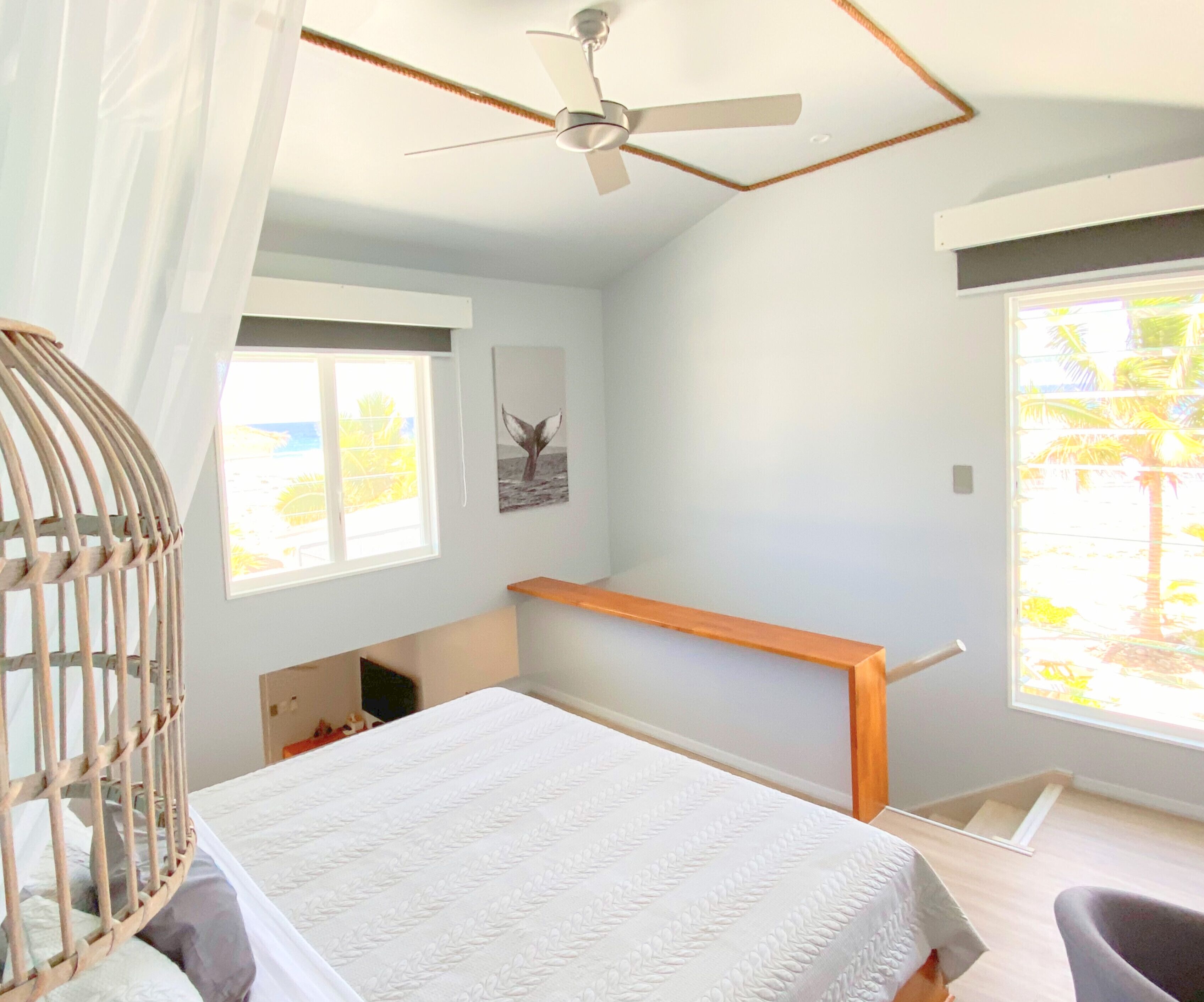 Un dormitorio con cama, ventilador de techo, ventana con vista a palmeras y estante de madera.