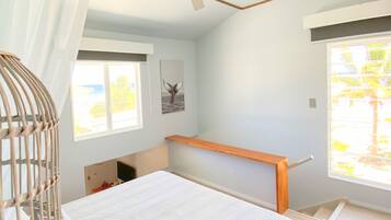Un dormitorio con cama, ventilador de techo, ventana con vista a palmeras y estante de madera.