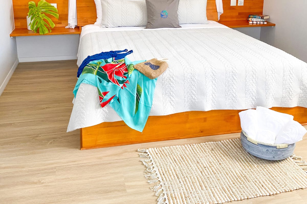 A bedroom with a wooden headboard, a large bed with white bedding, a bedside table with books, a hanging light fixture, and a round mirror on the wall.