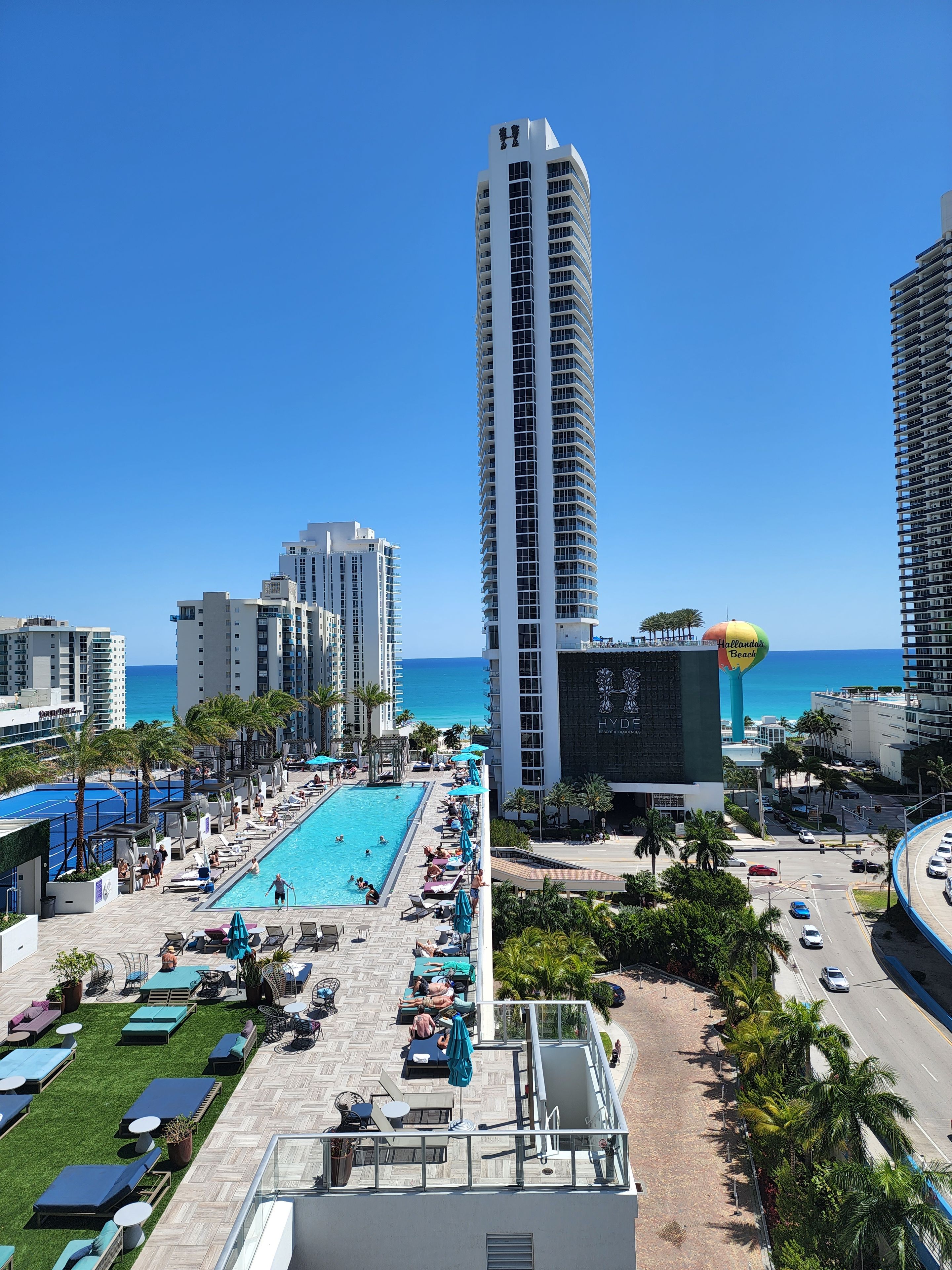 A high-rise hotel with a rooftop pool area, palm trees, and a view of the ocean.