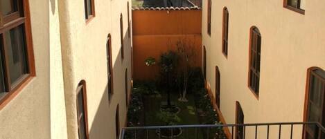 Un patio estrecho con un pequeño jardín, un balcón con barandilla de metal y edificios con ventanas de arco.