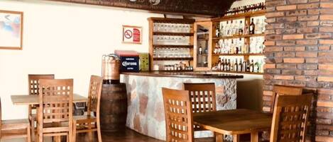 Zona de bar con pared de ladrillo, sillas de madera, mostrador y una variedad de botellas.