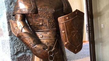 Una estatua de bronce de un caballero con armadura, sosteniendo un escudo, se encuentra en un pasillo con un muro de piedra y una puerta al fondo.