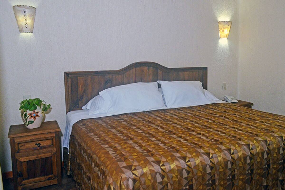 Ein Schlafzimmer mit einem hölzernen Kopfende, einer gemusterten Bettdecke, einem Nachttisch mit Pflanze und einem an der Wand befestigten Telefon.
