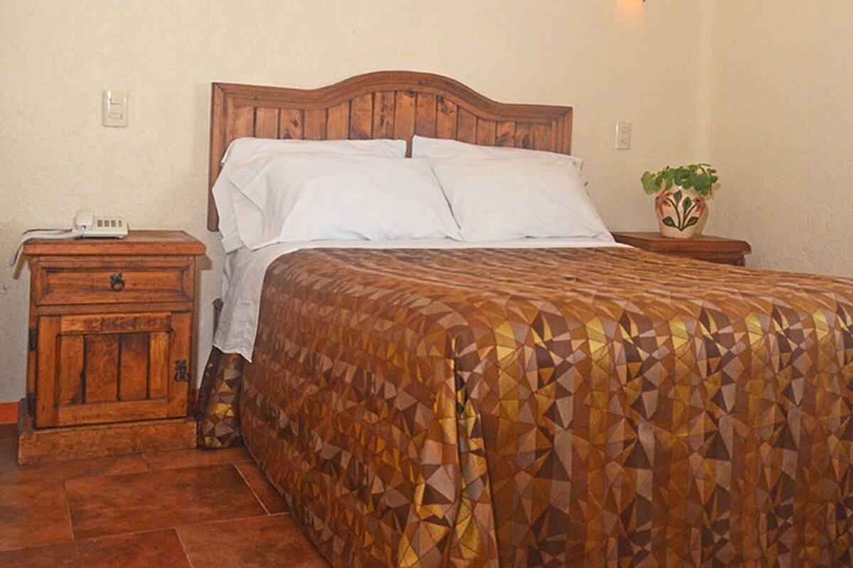 Ein Schlafzimmer mit einem Holz-Kopfteil, einer gemusterten Bettdecke, einem Nachttisch mit Telefon und einer Topfpflanze.