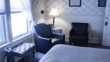Une pièce comprenant un lit, un fauteuil bleu, un fauteuil noir, une petite table avec une lampe et un banc.