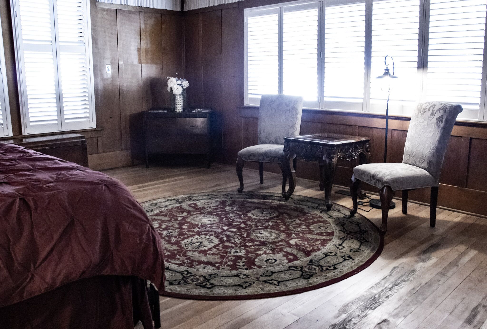 A room with a large bed, a round rug, two chairs, a small table, and a window with blinds.