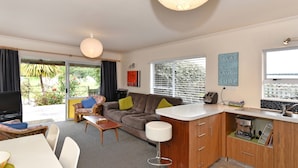 A modern living room with a sofa, a coffee table, and a dining area with a table and chairs. There is a kitchen area with a countertop and cabinets. - Blue Lagoon - Kaiteriteri Holiday Unit (Kaiteriteri)