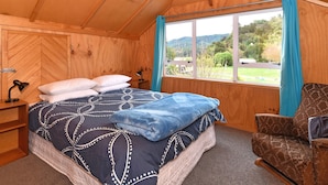 A wooden room with a bed, a chair, a desk, and a window with curtains. - Blue Lagoon - Kaiteriteri Holiday Unit (Kaiteriteri)