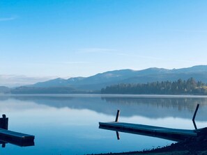 A calm lake with a dock extending into the water, surrounded by mountains and trees. - Lakefront condo w/ beach and seasonal heated pool (Mccall)