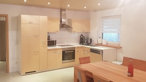 Une cuisine moderne avec des meubles en bois clair, un four à micro-ondes, une cuisinière et un lave-vaisselle. On y trouve une table à manger avec des chaises et une bouteille de vin posée sur la table.
