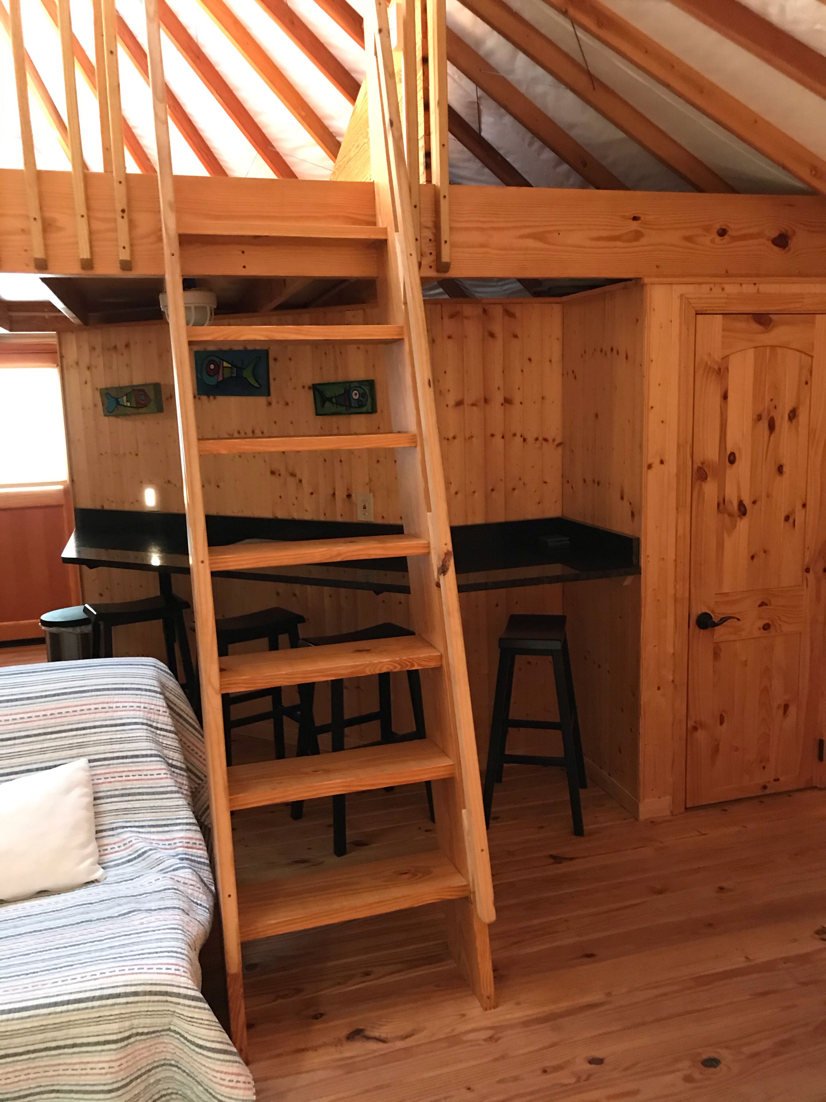 A loft-style bedroom with a wooden ladder leading to a lofted sleeping area. There is a desk and two stools in the room.