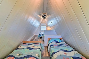 Une pièce étroite et inclinée, avec deux lits, un ventilateur de plafond et une petite table.