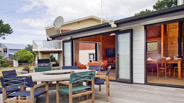 An outdoor deck with a dining table and chairs, a barbecue grill, and a large sliding glass door leading to a living area with a television. - Pretty in Pauanui - Pauanui Holiday Home (Pauanui)