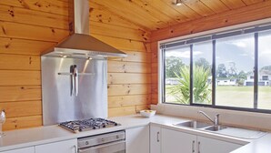 A modern kitchen with wooden walls, a stainless steel refrigerator, a gas stove, an oven, a sink, and a large window offering a view of the outdoors. - Pretty in Pauanui - Pauanui Holiday Home (Pauanui)
