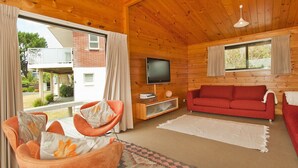 A wooden cabin interior with a red sofa, orange armchairs, a TV, and large windows. - Pretty in Pauanui - Pauanui Holiday Home (Pauanui)