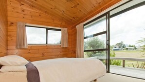 A wooden-paneled bedroom with a bed, two windows, and a view of the outdoors. - Pretty in Pauanui - Pauanui Holiday Home (Pauanui)