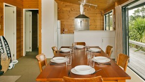 A wooden dining room with a large table set for six, a kitchen area, and a view of greenery outside. - Pretty in Pauanui - Pauanui Holiday Home (Pauanui)