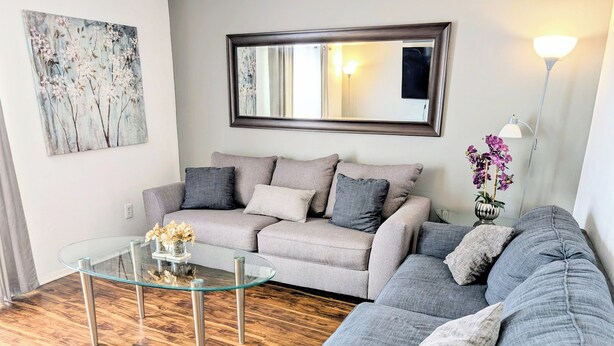 Uma sala de estar com um sofá cinzento, uma mesa de centro de vidro e um quadro na parede.