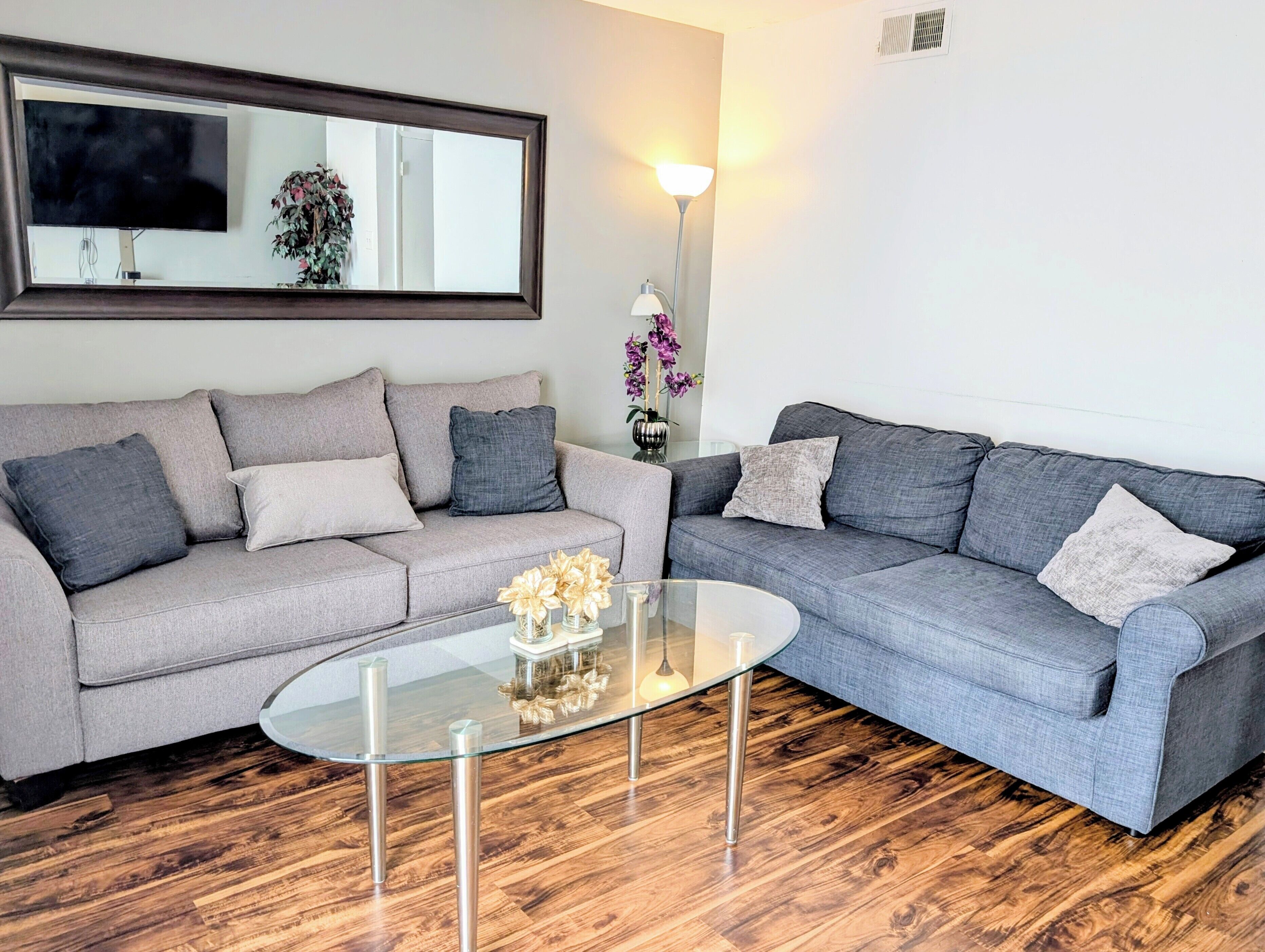A modern living room with two grey sofas, a glass coffee table, and a large mirror above a floral arrangement.