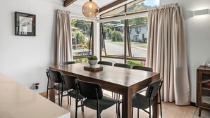 A dining area with a wooden table, black chairs, a hanging light fixture, and a view of a street and houses through large windows.