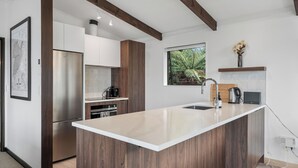 A modern kitchen with a central island, stainless steel appliances, wooden beams, and a window with a view of greenery.