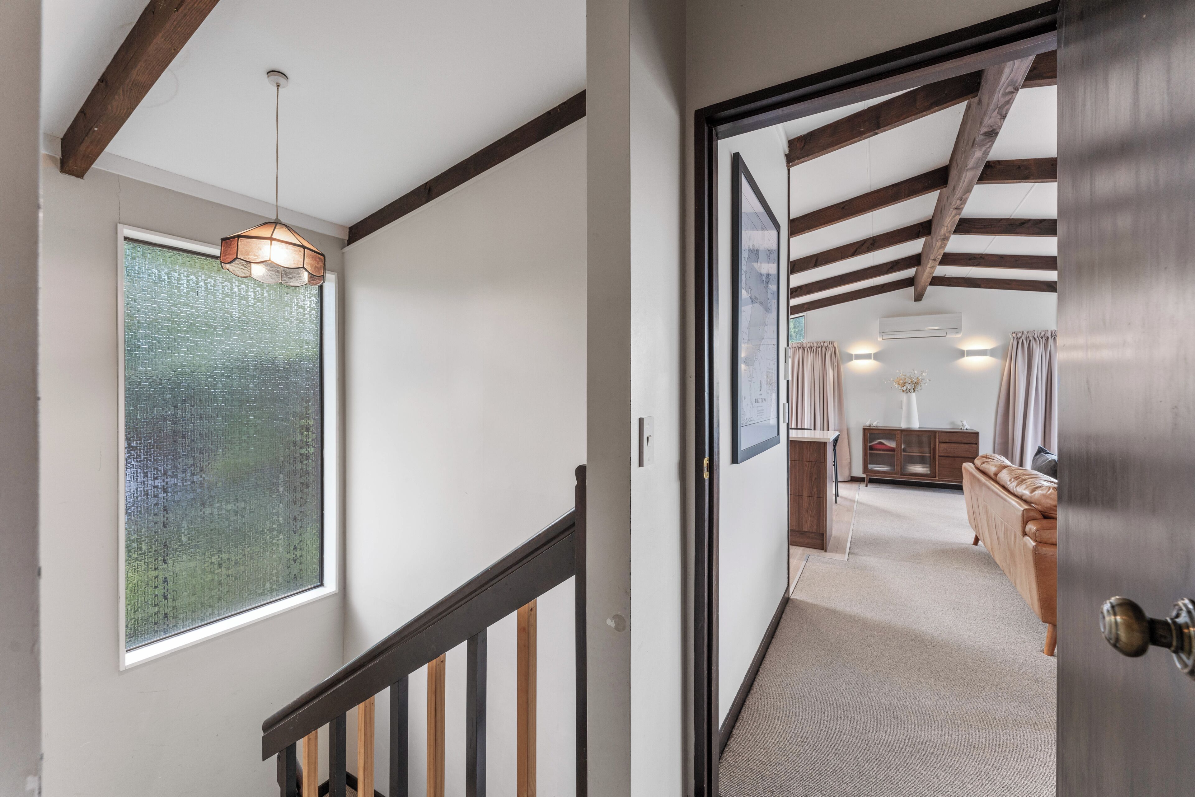 A hallway with a staircase, a wooden ceiling, a hanging light fixture, a window, and a door leading to a room with a sofa and a cabinet.