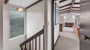 A hallway with a staircase, a wooden ceiling, a hanging light fixture, a window, and a door leading to a room with a sofa and a cabinet.