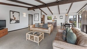 A spacious living area with a sofa, coffee table, and a television. There is a dining area with a table and chairs, and a kitchen in the background.