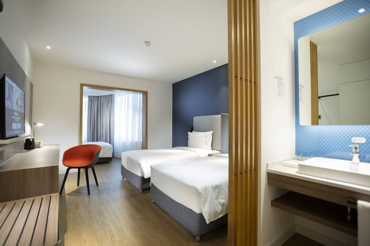 Habitación de hotel con dos camas, un escritorio, una silla, un baño con espejo y un televisor.