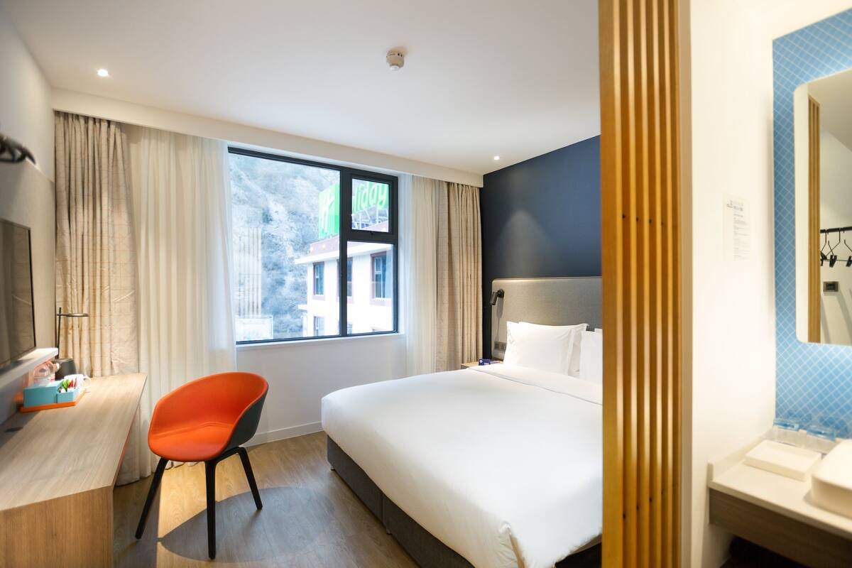 Habitación de hotel con cama, escritorio, silla, ventana con cortinas y baño con espejo y lavabo.