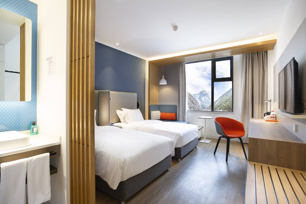 Habitación de hotel con dos camas, un escritorio, una silla, un televisor y una ventana con vista a la montaña.