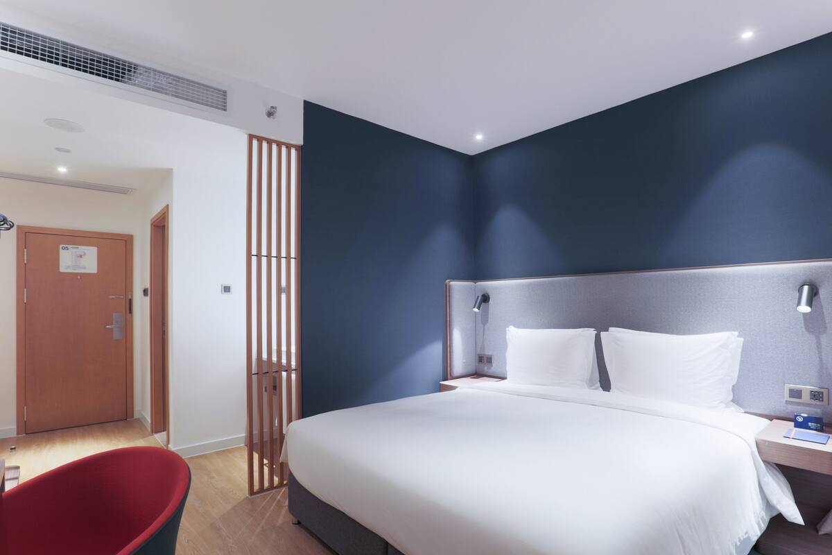 Una habitación de hotel moderna con una cama grande, una mesita de noche con lámpara y una silla roja.