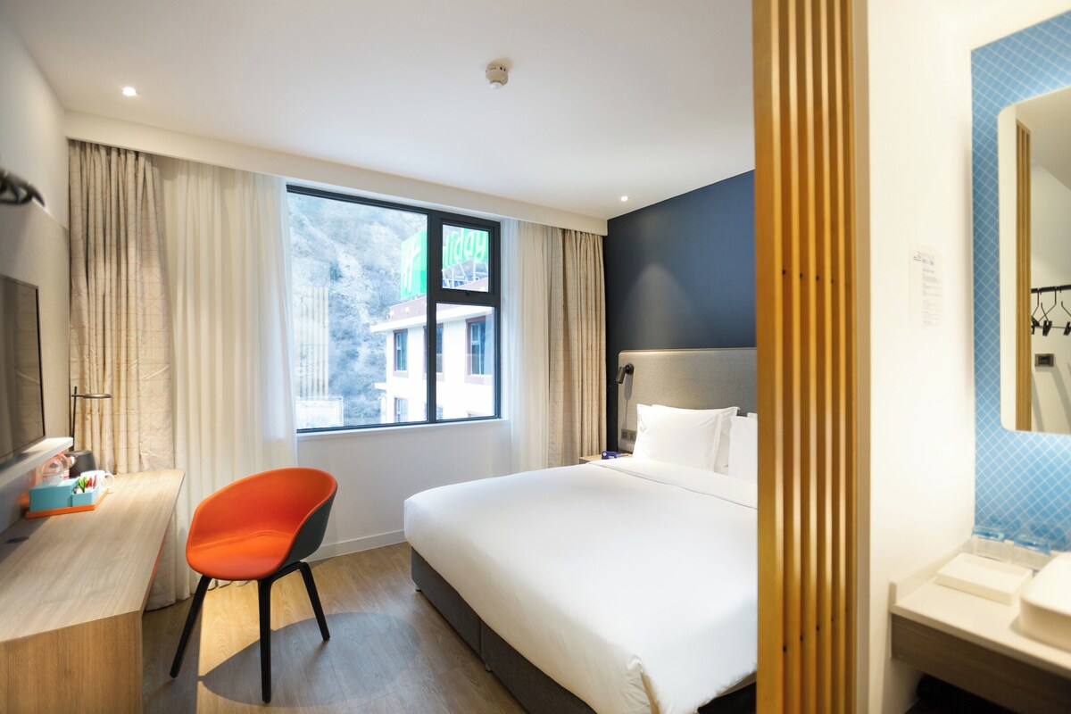 Une chambre d’hôtel comprenant un lit, un bureau, une chaise, une fenêtre avec des rideaux et une salle de bain avec un miroir et un lavabo.