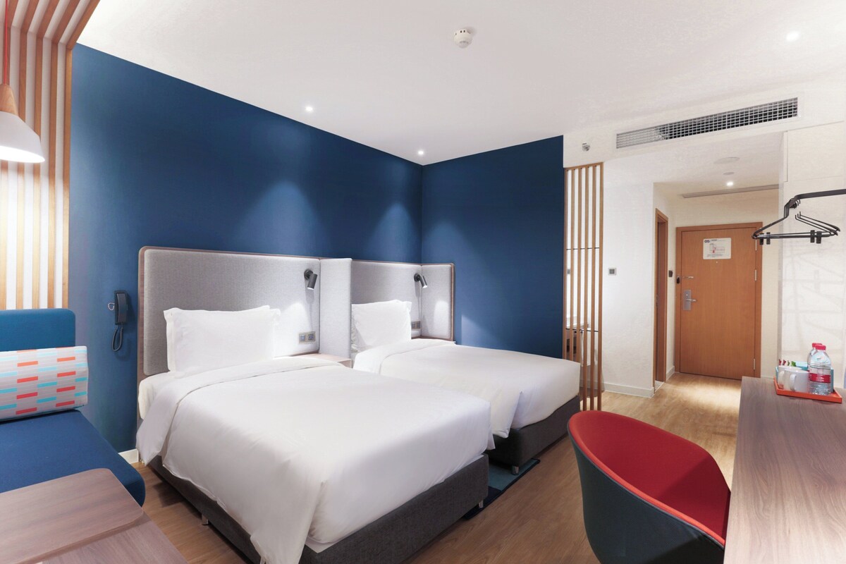 Une chambre d’hôtel avec deux lits, un bureau, une chaise et une salle de bain visible à travers une porte ouverte.