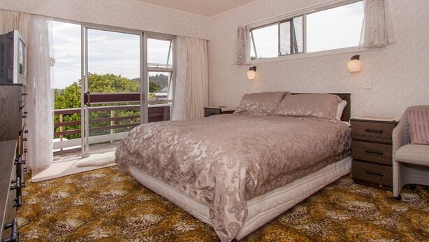 A bedroom with a large bed, a dresser, a chair, and a TV. There is a balcony with a view of greenery. - Holiday Haven - Whitianga Bach (Whitianga)